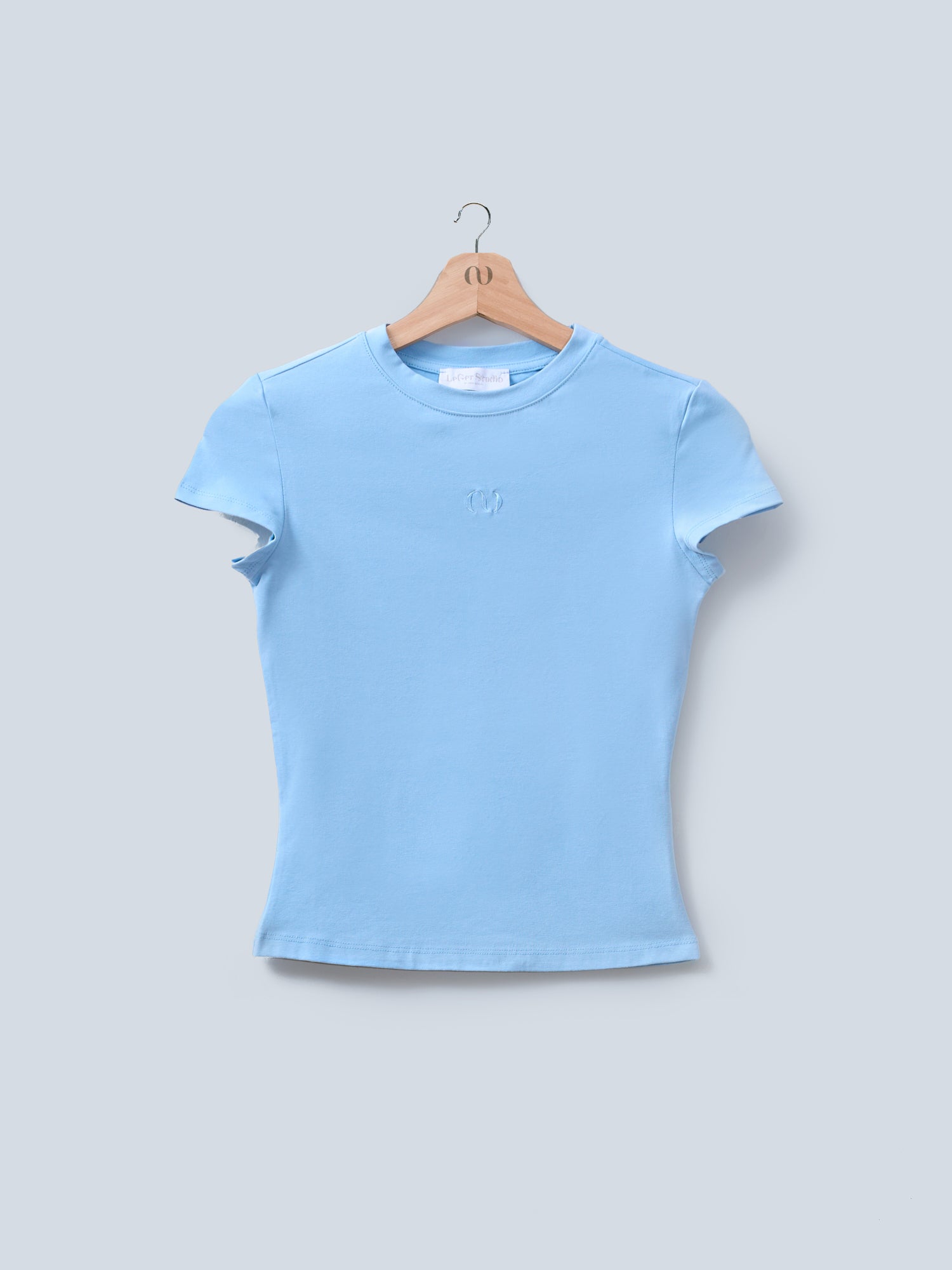 Das Franka T-Shirt von LeGer by Lena Gercke ist ein hellblaues, eng anliegendes, kurzärmliges Oberteil, das auf einem Holzbügel präsentiert wird. Dieses stilvolle Kurzarm-Design verfügt über ein dezent gesticktes Logo auf der Brust.