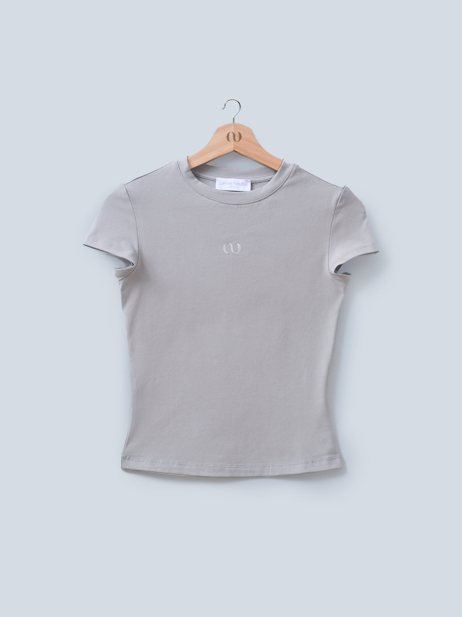 Das T-Shirt „Franka“ von LeGer by Lena Gercke, mit dezenter Logostickerei auf der Brust, in Grau mit kurzen Ärmeln, hängt elegant auf einem Holzbügel vor hellgrauem Untergrund.