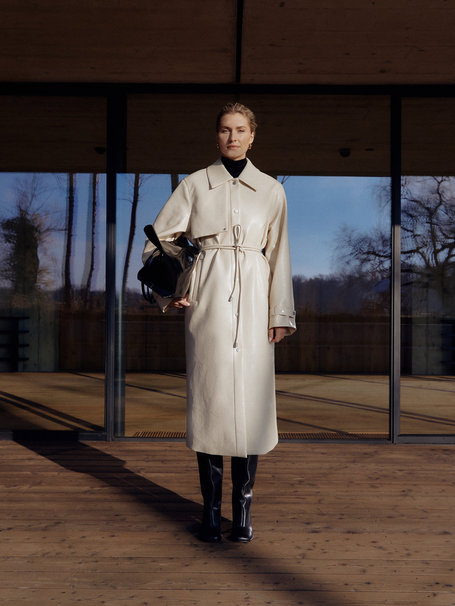 In einer sonnigen Umgebung im Freien steht jemand auf einem Holzdeck vor einem Glasgebäude und trägt den Birka Coat von LeGer by Lena Gercke. Der beige Trenchcoat mit eleganter langer Passform wird mit einem schwarzen Rollkragenpullover und einer schwarzen Hose kombiniert, während die Person eine schwarze Tasche trägt, eingerahmt von Bäumen im Hintergrund.