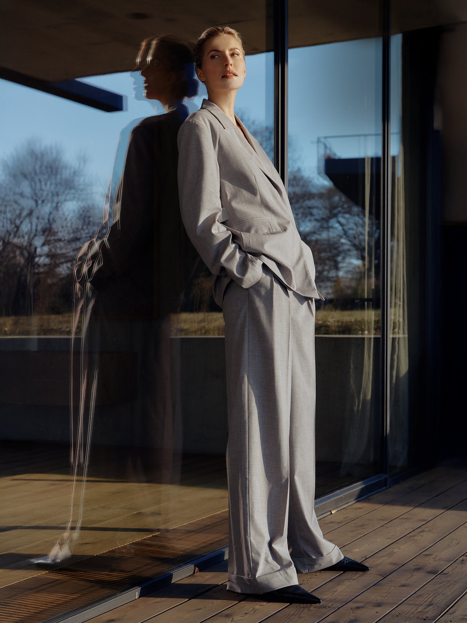 Eine Person in locker sitzenden Jale Pants Tall von LeGer von Lena Gercke, hellgrau mit Bügelfalten, steht mit den Händen in den Taschen vor einer Glaswand, die ihr Bild reflektiert. Im Hintergrund sind blattlose Bäume und Grasfelder unter einem klaren Himmel zu sehen.